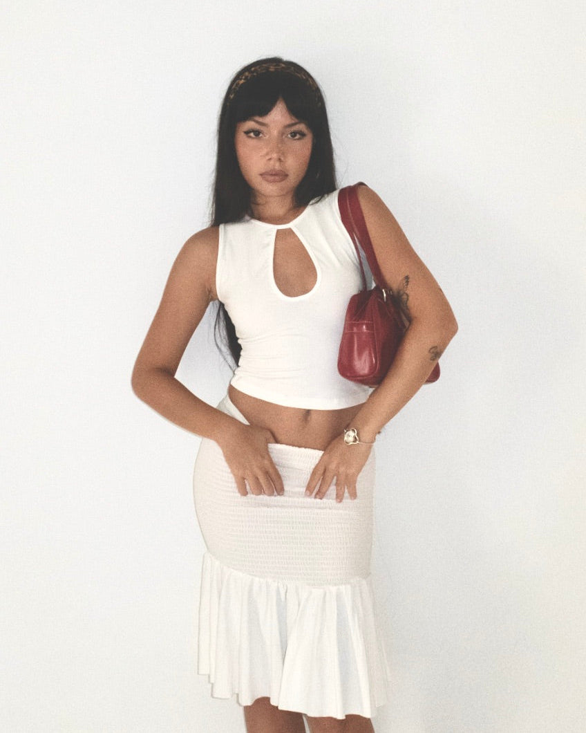 Woman wearing a white sleeveless top and skirt with a red handbag on a plain background
